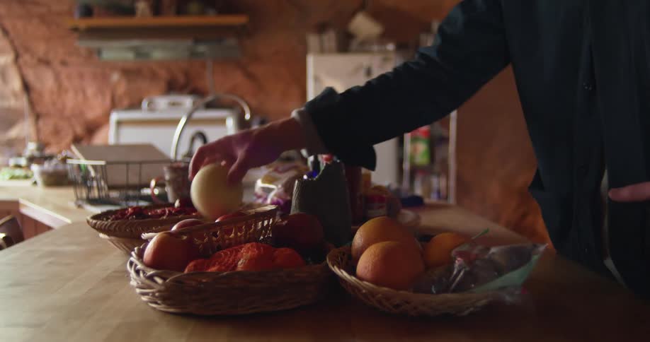 A man at home picks up an apple and then an orange out of baskets on his counter