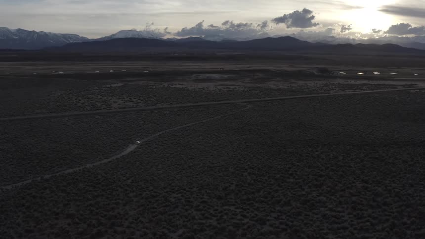A vehicle travels along a dirt road through a shrubland in the evening