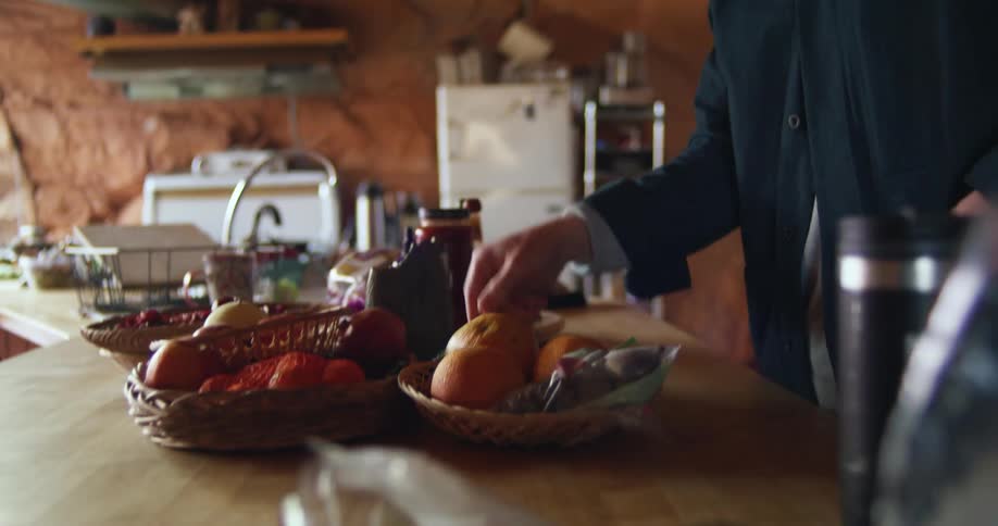 A man in a cave room smelling a fruit with a wall and refrigerator in the background