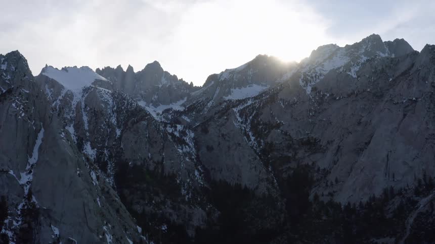 A mountain range with snow with the sun the sky and clouds in the background
