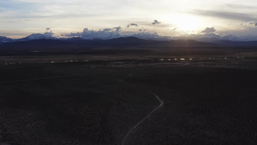 Winding road leads through scrub covered plains to town backed by mountains under cloudy sky
