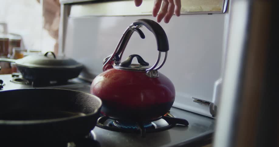 A hand picks up a kettle and places it on a gas burner on a stove