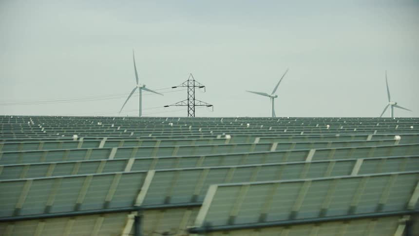 Passing row after row of solar panels with rotating wind turbines and high transmission power lines in the background in this energy intensive shot