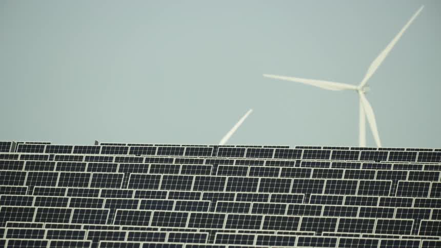 Row after row of solar panels in a solar farm with rotating blades of a wind turbine are seen in this dynamic and awesome renewable energy shot