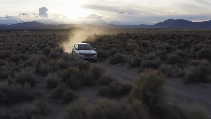 Suv drives along dirt road through scrub kicking up dust with mountains on horizon