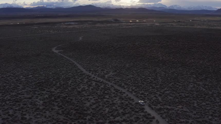 A vehicle driving on a dirt road in the desert with a mountain range the sky and clouds in the background