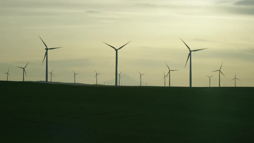 Many silhouetted wind turbines generating clean renewable electricity as the camera passes by the rural scene