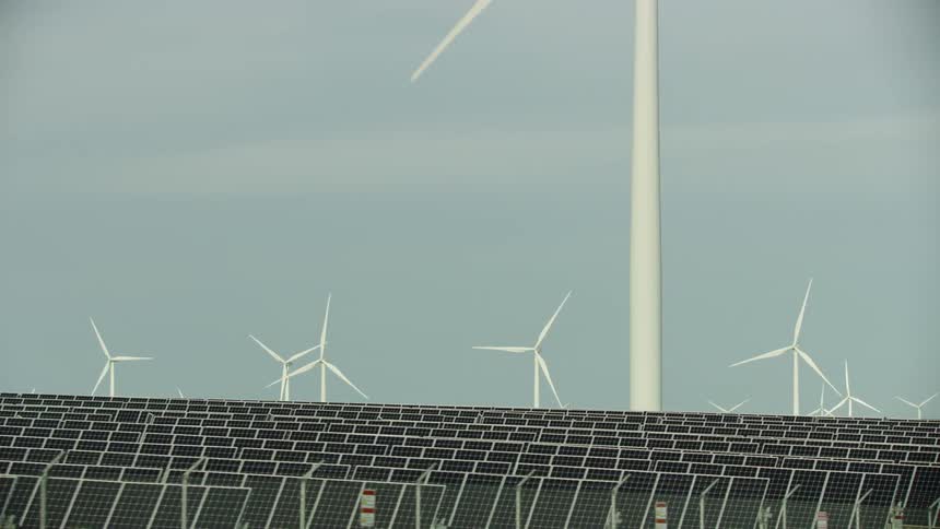 Spinning wind turbines loom over a vast array of solar panels on an energy farm during the day showing two different kinds of renewable energy generation in one shot