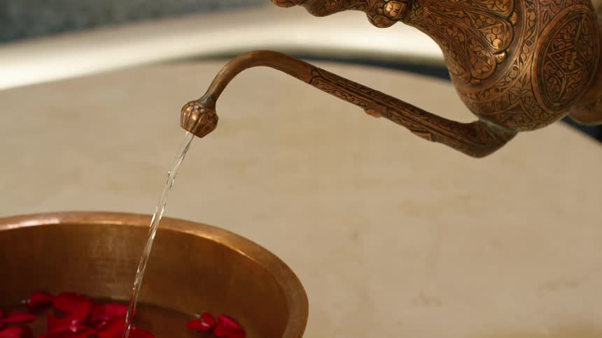 Person pours water from ornate pitcher into metal bowl of floating rose petals