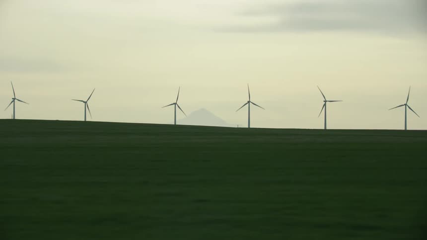 Zoom out reveal from a large mountain revealing many wind turbines generating renewable electricity with a green pasture foreground as seen from the Point of View of a driving car