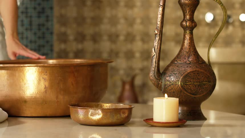 A woman in a towel is walking around a table of items which include a candle and a gold bowl