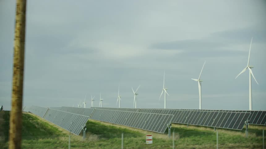 Passing row after row of solar panels on a solar farm as the camera pans left from wind turbines to the solar farm alone visually communicating renewable energy creation