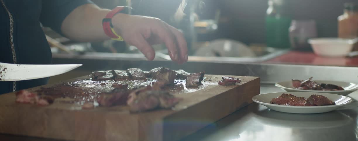 A person in a room holding a knife touching cooked meat with smoke with a sink and faucet in the background