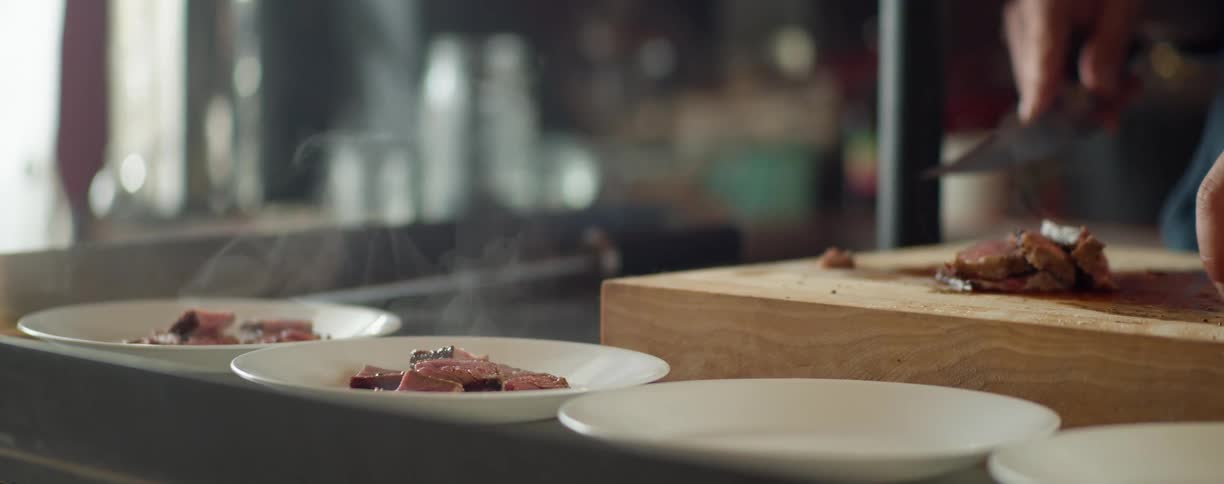 A man places slices of steaming hot meat onto plates as his friend puts a beer down