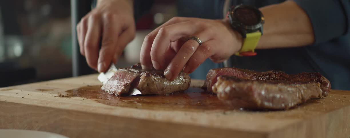 A person with a watch and ring cutting meat with a knife on a cutting board
