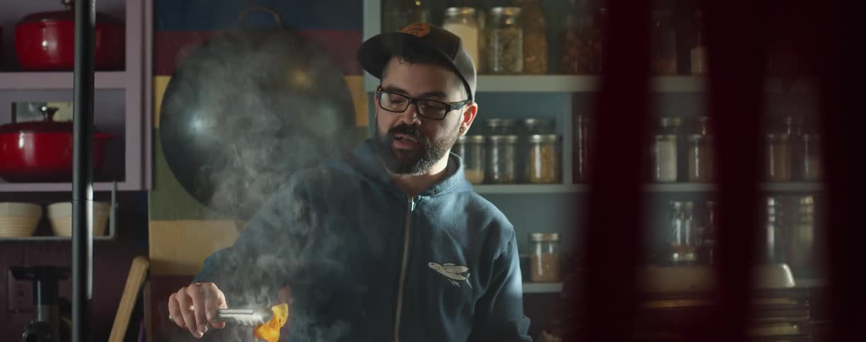 A man in a room with a hat and glasses cooking food with shelves in the background