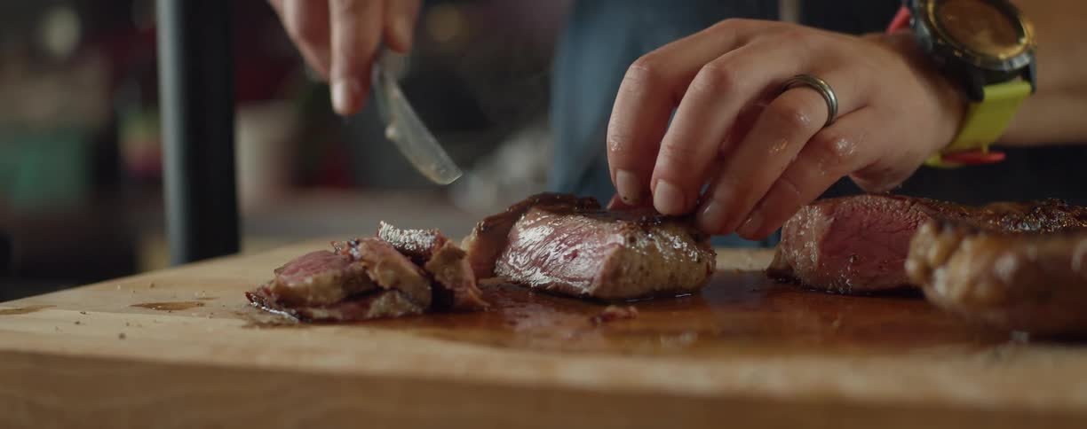 A person with a watch and ring cutting meat with a knife on a cutting board