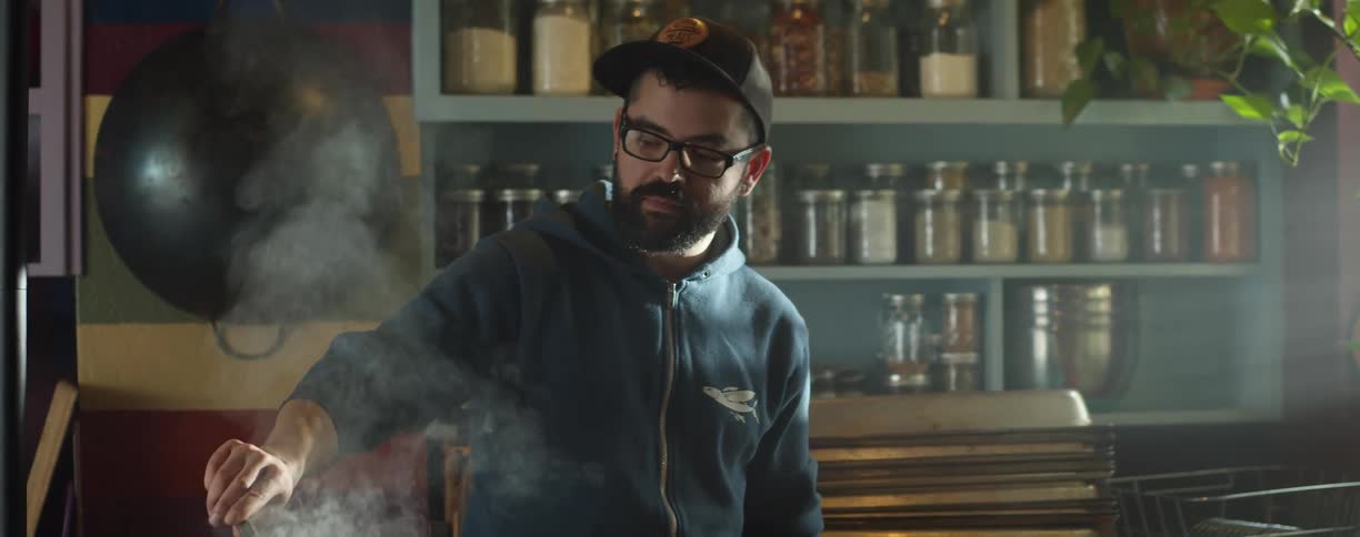 A man in a room with a hat and glasses cooking food with shelves in the background