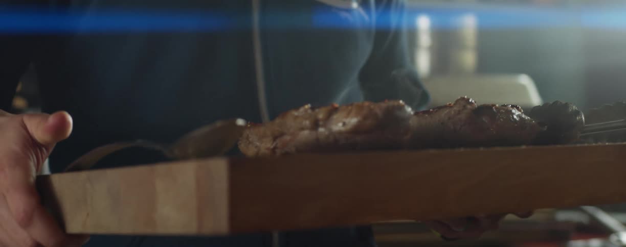 A person in a kitchen picking up a cutting board with cooked meat with a wall and window in the background