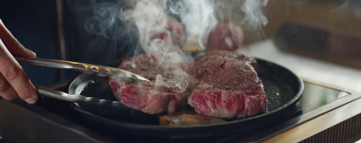 A person in a room cooking meat on a pan with smoke