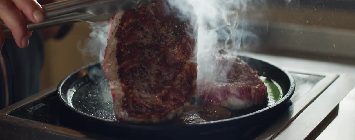 A man uses a pair of tongs to flip a steak that is being grilled in a circle pan on a small stove top