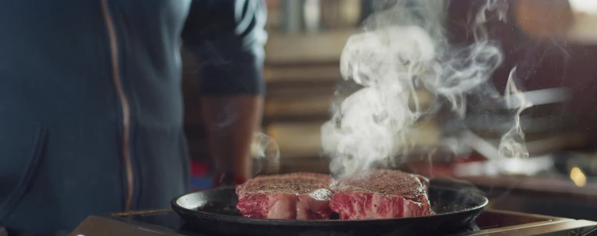 A person cooking meat on a pan with smoke with shelves in the background