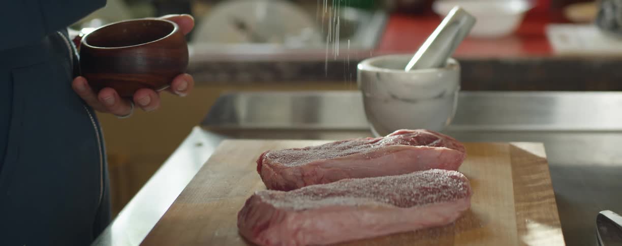 A person in a kitchen sprinkling salt on meat with a wall and window in the background