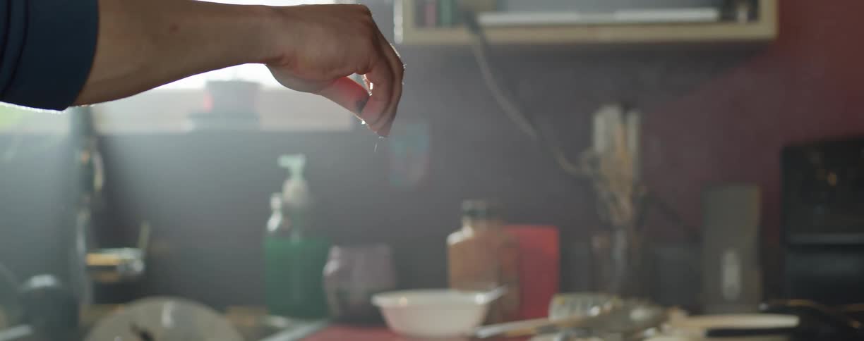 A person sprinkling salt in a kitchen with a wall and window in the background