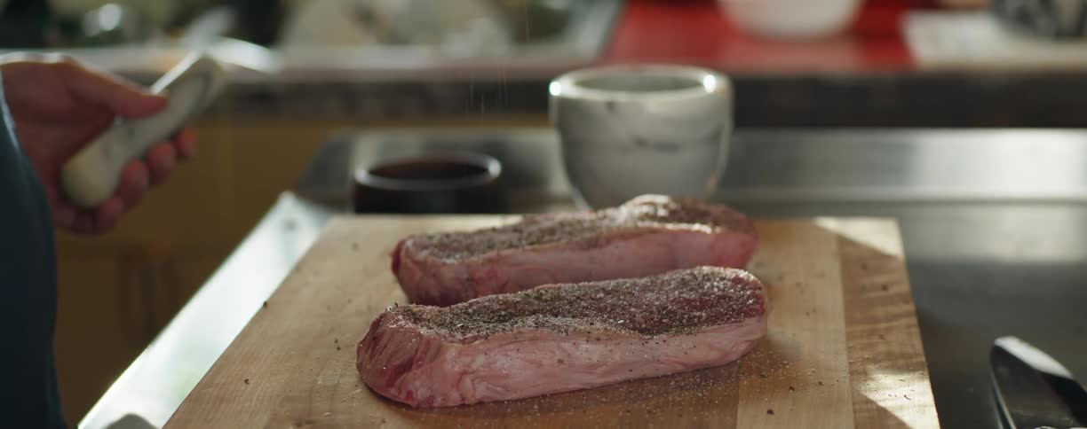 Person holding pestle seasons thick steaks sitting on wooden board