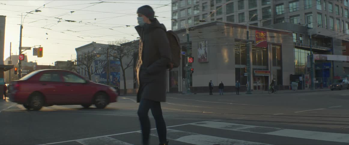 A person with a hat a mask and a backpack crossing a city street with buildings the sky and clouds in the background