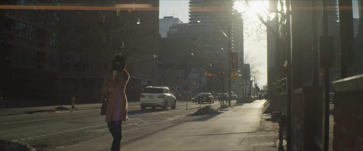 A woman with a mask walking on a city pavement with vehicles buildings trees the sky and clouds in the background