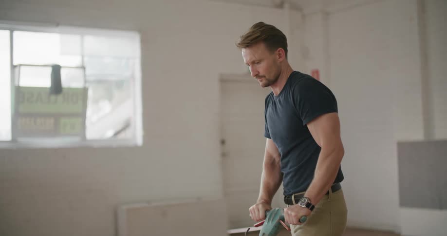 A man wipes his forehead with his forearm as he uses a buffing machine to buff the wooden floor of an empty room
