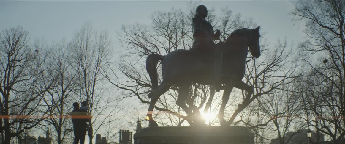 Father picks up child near statue and are also silhouette by sun shining through city