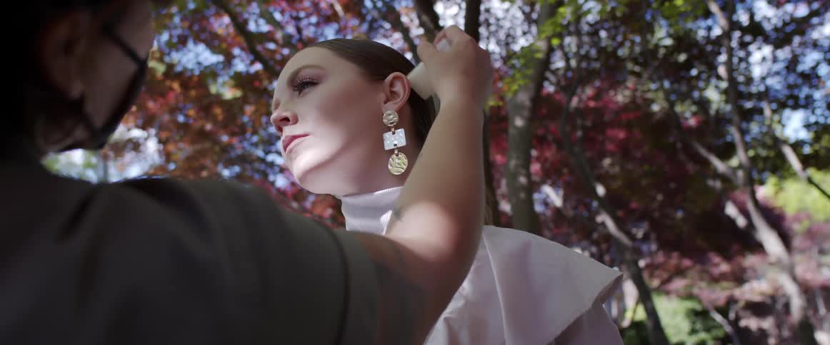 A woman in a medical mask applies makeup to a posing model outdoors during the day