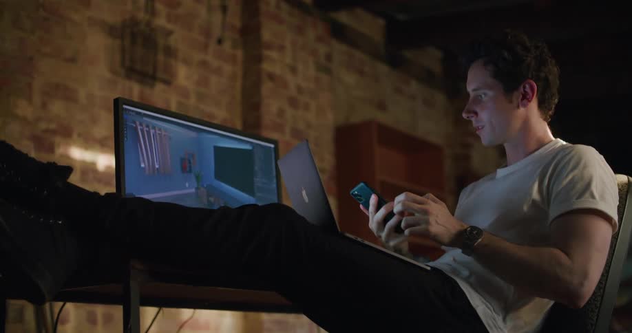 A young man sitting at a desk speaks on his smartphone before putting it away and typing on his laptop