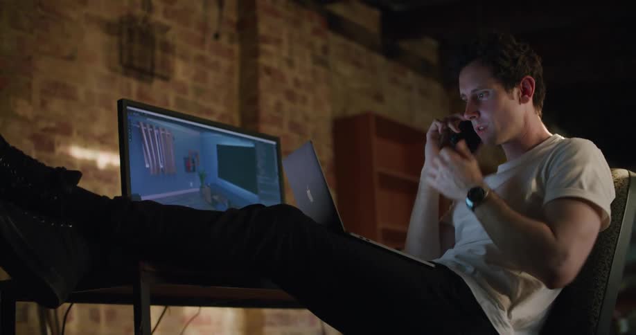 A man in a room sitting at a desk with a computer talking on a smart phone and typing on a laptop