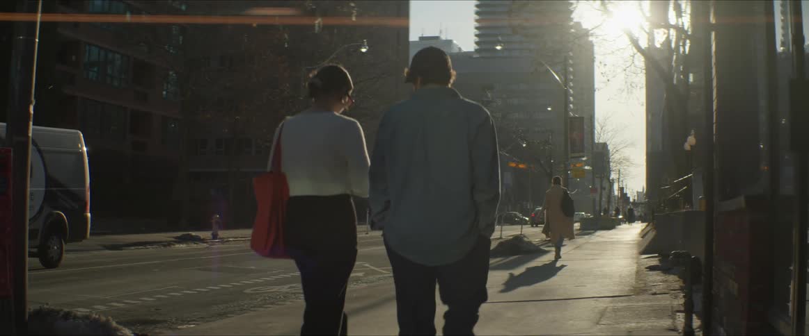 People walk on the sidewalk of a city street with melting snow at the curb and sun shining
