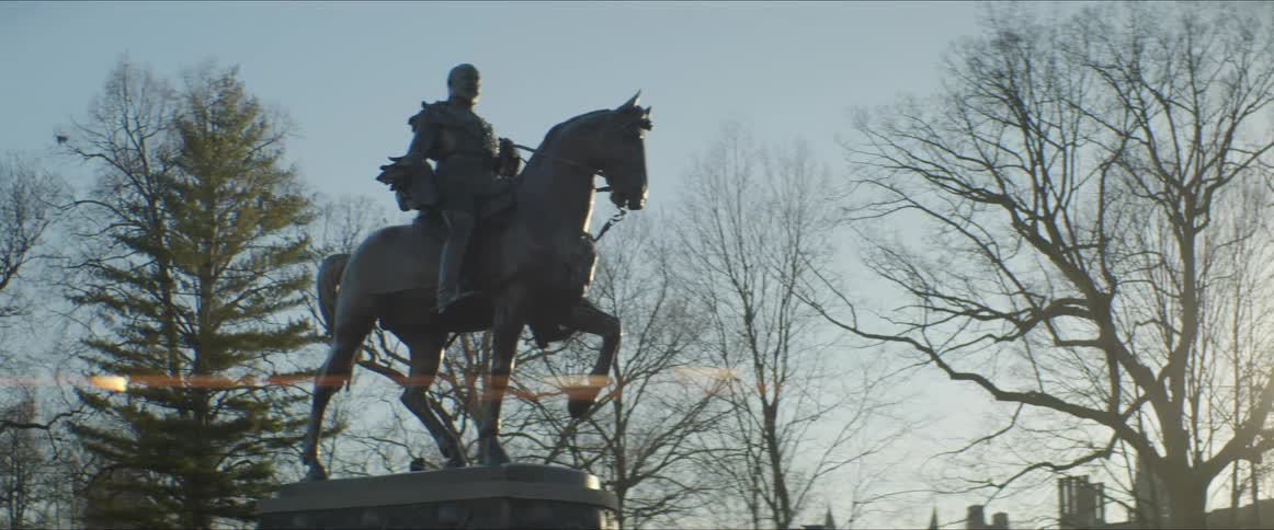 A statue of a man on a horse stands in a park with buildings visible through the trees