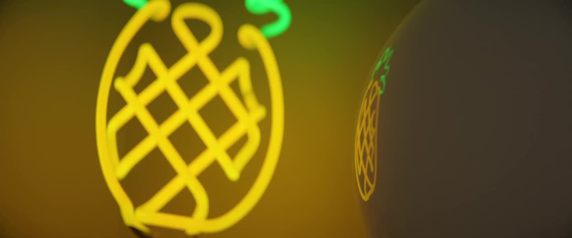 A pineapple shaped neon sign on the wall is reflected off a nearby round lampshade
