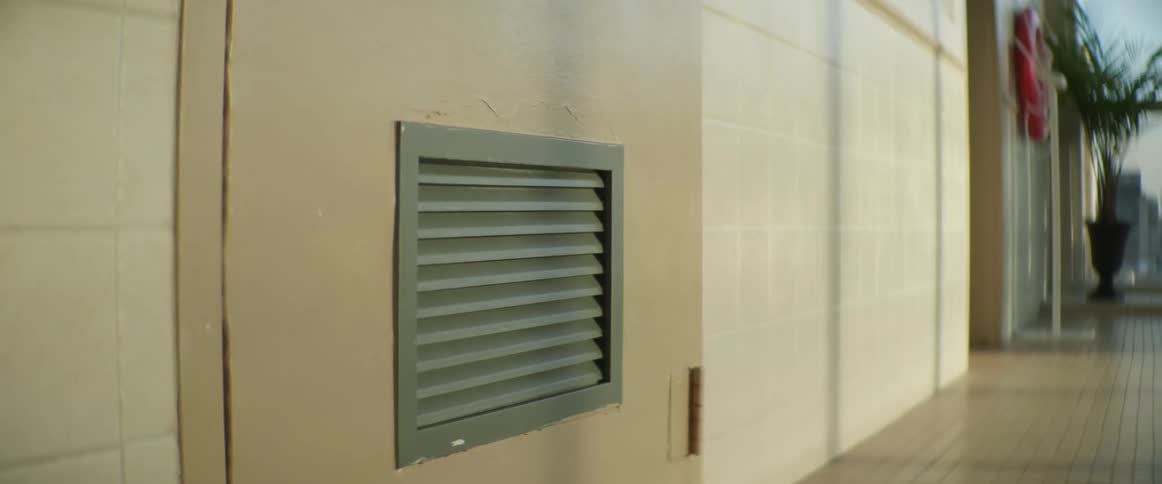 A silver vent on a door on the side of a building outdoors with a reflection of water on the wall