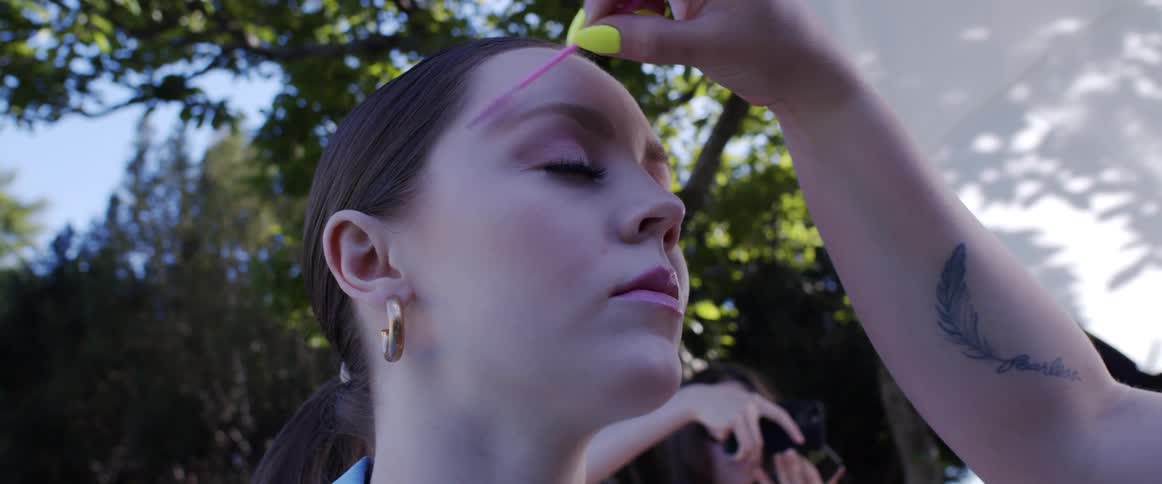 A woman standing as another woman puts makeup on her with trees the sky and clouds in the background
