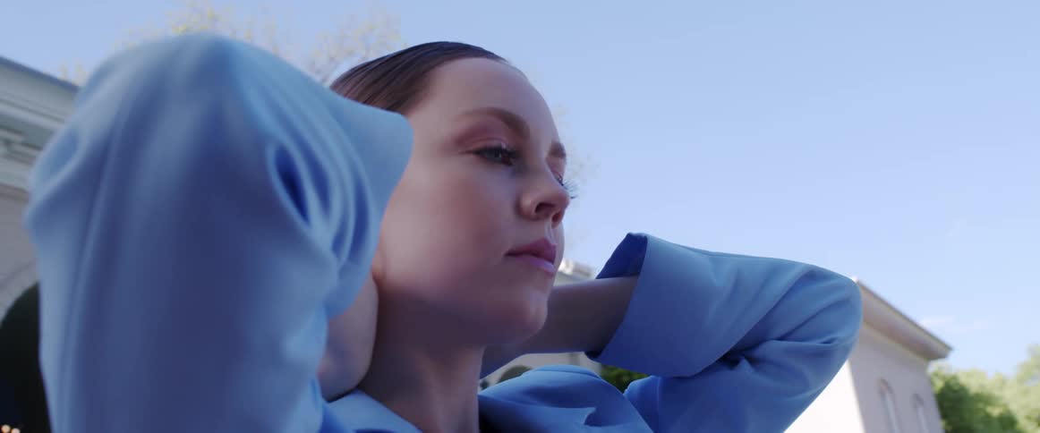 A model poses in a blue suit jacket with her hands behind her head in front of a white building with large arched entrances