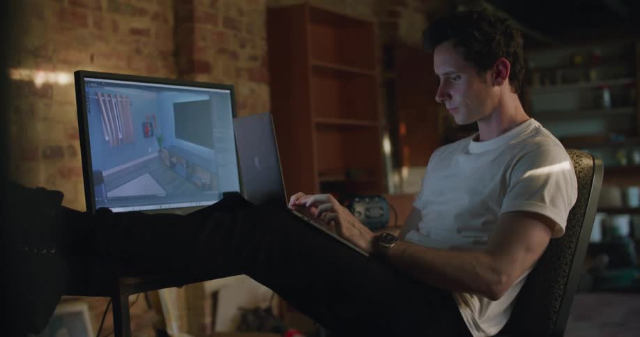 A man stretches his neck as he types on his laptop with his feet up at a desk indoors during the day