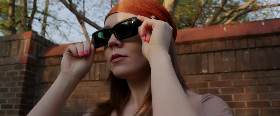 A woman wearing an orange head scarf adjusts her black sunglasses as she stands in front of a brick wall