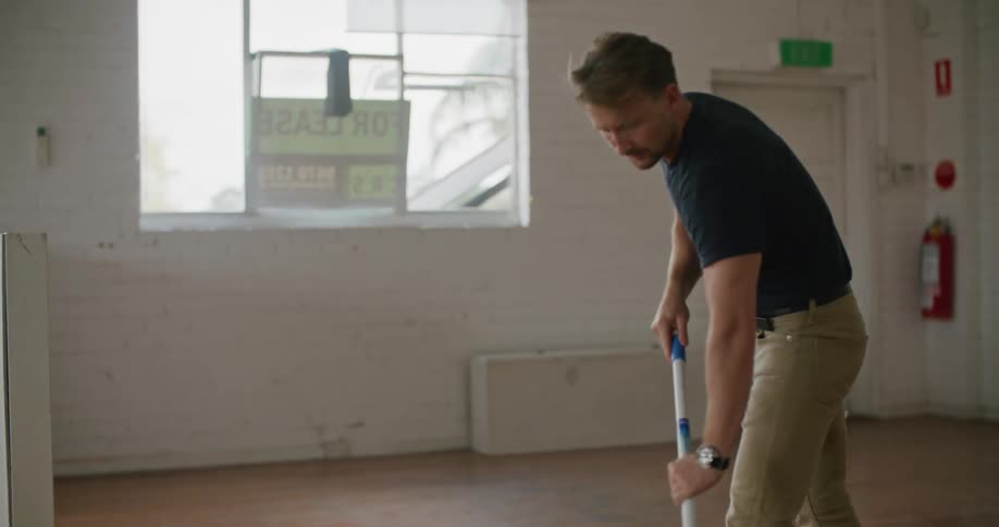 A man mops the floor of an empty room for a moment before stopping to rest