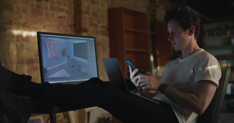 A man in a room sitting on a desk with a laptop and computer