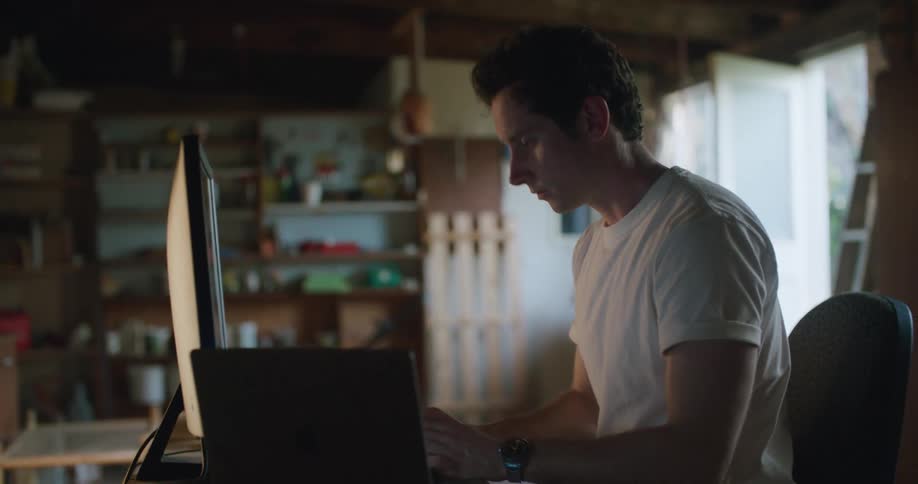 A man with a smart phone sitting at a desk with a computer and laptop