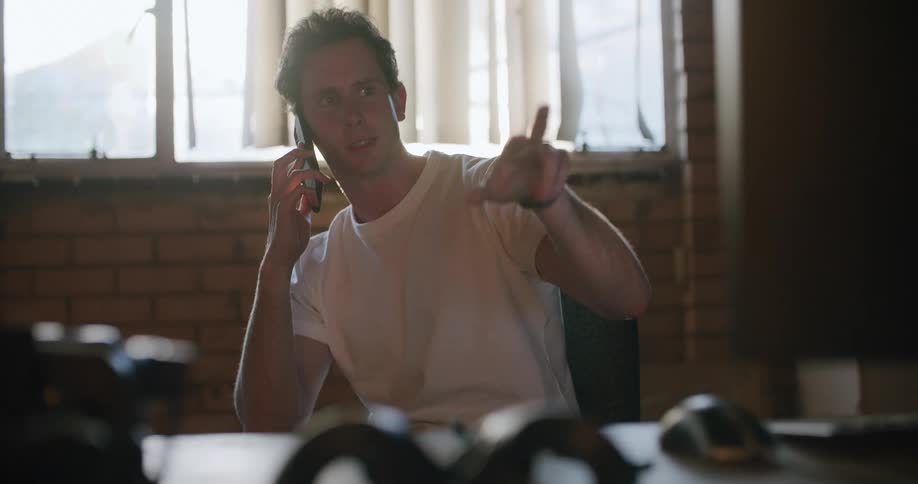 A man in a room sitting on a chair talking on a smart phone with a wall and window in the background