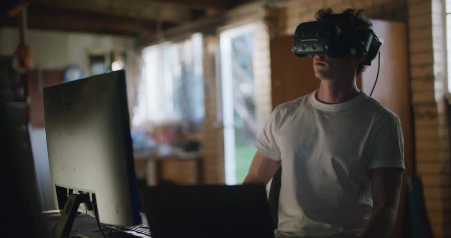 A man looks up turns his head and adjust his vr glasses as he sits in front of a computer
