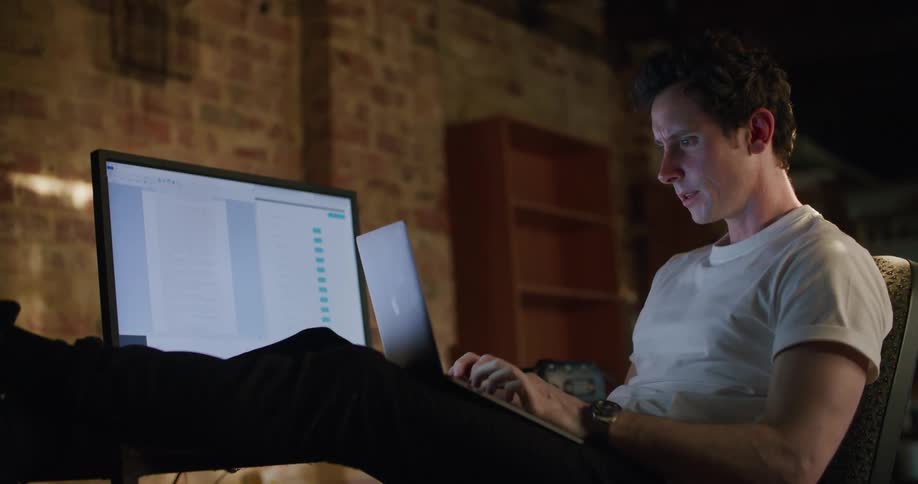 A man sits with his legs on his desk and a laptop on his lap as he leans back contemplating